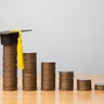 Mortarboard or square academic cap and reduce stack coins chart on wooden table with white wall background. 