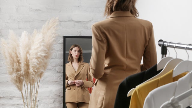 Confident woman with blond hair standing in front of mirror in fashionable boutique looking at reflection 