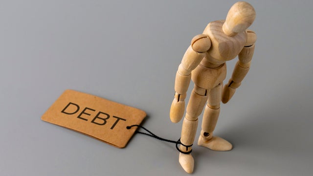 A wooden figure stands with one ankle tied by a string to a tag labeled DEBT. 