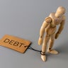 A wooden figure stands with one ankle tied by a string to a tag labeled DEBT. 