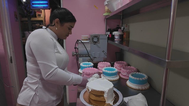 Ciddianys Rodriguez ices a cake in her bakery, pink and blue cakes are on the table nearby 
