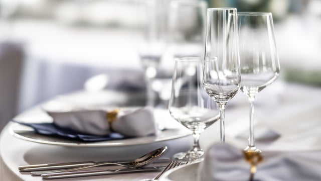 The photo shows an elegant table setting at a formal event, featuring polished silverware and clean white linens with blue accents. Several crystal-clear wine and champagne glasses stand in the center, catching the light against a soft, blurred background 